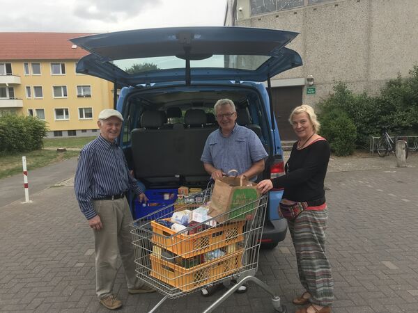Zwei Männer, eine Frau vor einem geöffneten Kofferraum mit einem einkaufswagen voller Lebensmittel